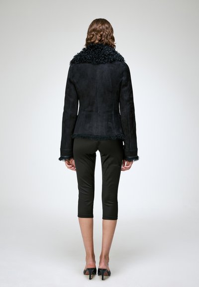 Femme debout, de dos, portant une veste en shearling noire, un pantalon court noir et des chaussures à talons noires sur un fond blanc uni.