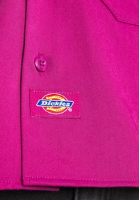 Camicia magenta con un bottone sul colletto e un colorato patch con il logo Dickies cucito nella zona della tasca. Tessuto liscio e testurizzato.