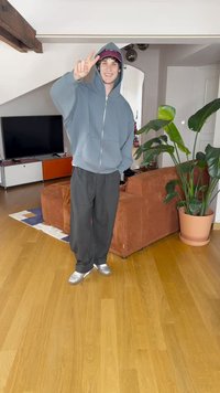 Joven con una sudadera azul y gorra sonríe, mostrando un signo de paz en una sala de estar con un sofá marrón, una planta grande y un televisor.