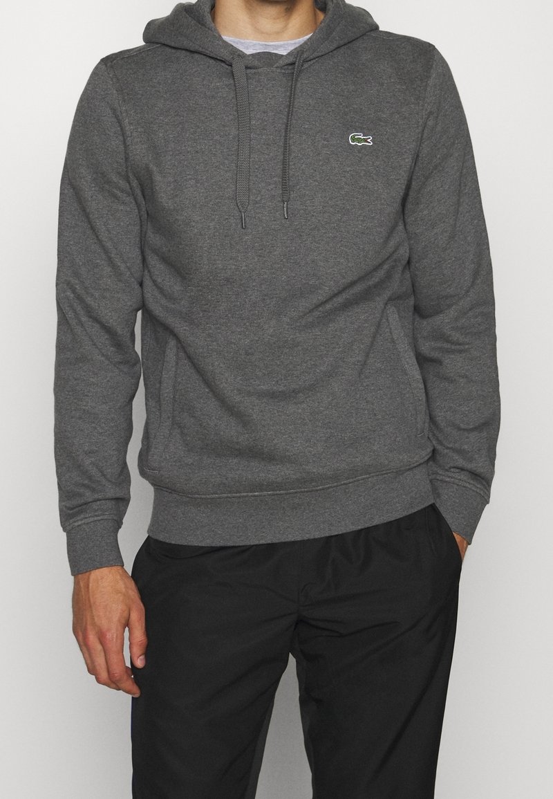 Homme portant un sweat à capuche gris foncé avec des cordons et un petit logo crocodile vert sur la poitrine, associé à un pantalon noir.