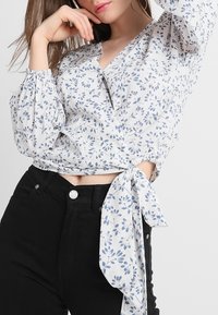 Blouse florale en tissu léger avec imprimé botanique bleu, décolleté en V, manches bouffantes et taille nouée, associée à un pantalon noir taille haute.