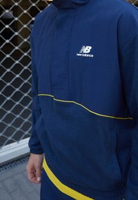 Tmavě modrá bunda s límcem ze syntetického materiálu se žlutou linkou, s vysokým límcem a logem New Balance v bílé barvě.