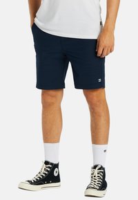 Navy blue shorts made of cotton blend, with a relaxed fit, side pockets, and a small logo detail near the hem; paired with black high-top sneakers.