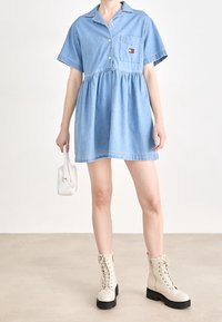 Ljust blå denimskjolklänning med korta ärmar, knäppning framtill och draperad midja. Har en bröstficka och är kombinerad med beige kängor.