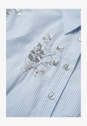 Lichtblauw-wit gestreept overhemd met zilveren pailletten bloemenborduursel bij de knopen en kraag.