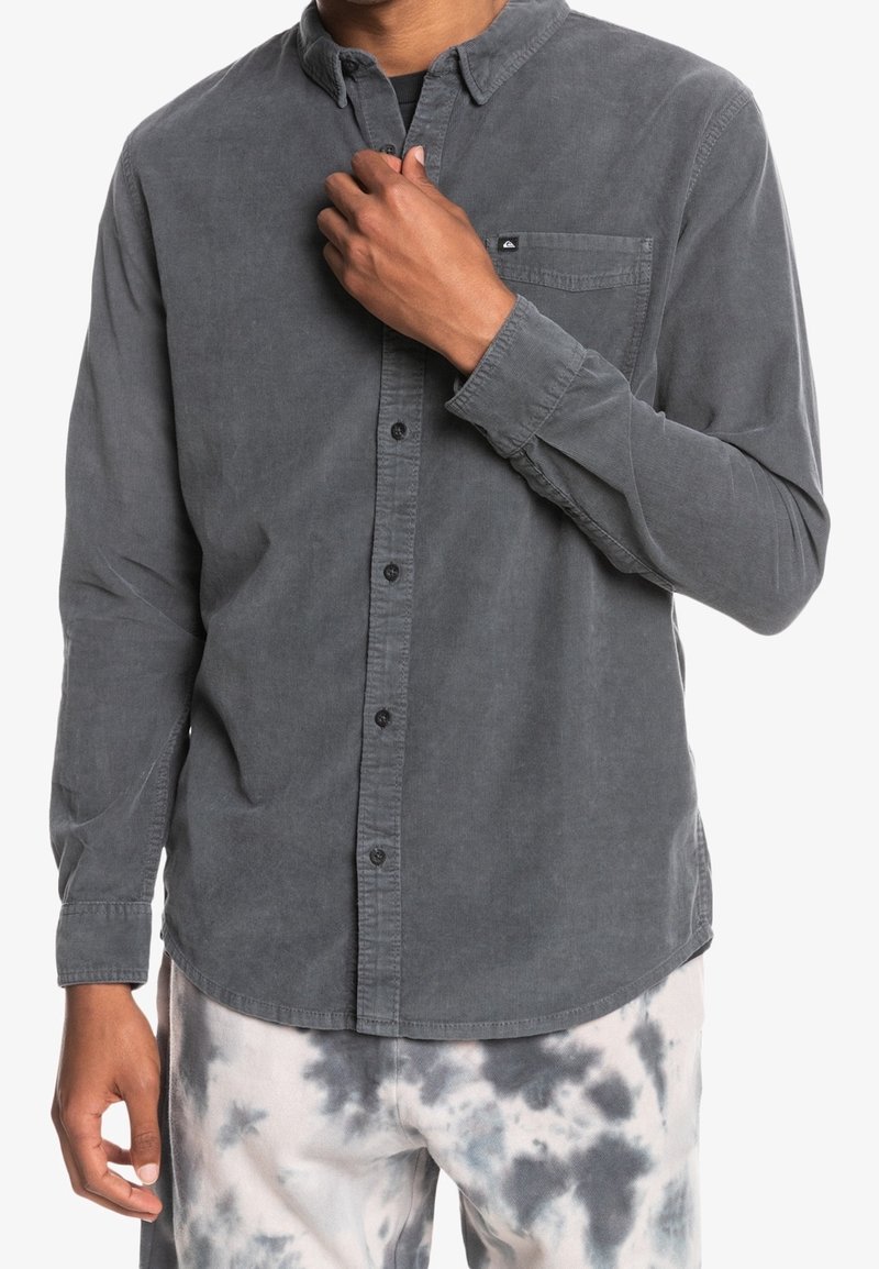 Szara koszula z kordurowego materiału z kołnierzykiem zapinanym na guziki, przednią kieszenią i czarnymi guzikami, noszona z spodniami w wzór tie-dye.