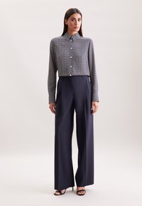 Camicia button-up con un motivo geometrico, abbinata a pantaloni a vita alta e gamba larga in un blu navy scuro. Tacchi neri con cinghie completano l'outfit.