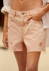 Shorts de mezclilla rosa con bordado floral, dobladillo deshilachado, diseño de cinco bolsillos, cierre de botón metálico y presillas para cinturón. Textura suave con costuras visibles.