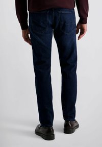 Man in donkere blauwe jeans, bruine schoenen en een kastanjebruine lange mouwen shirt, staande met zijn rug naar de camera op een effen lichte achtergrond.