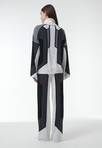 Vrouw met lang bruin haar, gekleed in een zwart, wit en grijs geometrisch patroon turtleneck sweater en wijde broek, met haar gezicht naar de andere kant.