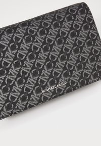 Černá textilní peněženka s geometrickým vzorem střídajících se písmen "MK." Stříbrné logo vyražené v pravém dolním rohu.