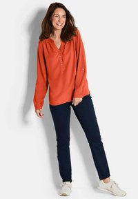 Orange Langarmbluse mit einer Knopfleiste und drei Knöpfen, kombiniert mit dunkelblauen Slim-Fit-Hosen und hellen Sneakers.