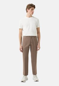 Roomkleurig T-shirt met korte mouwen gecombineerd met lichtbruine, op maat gemaakte broek en witte sportieve schoenen, staand tegen een effen achtergrond.