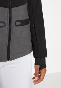 Czarna i szara kurtka z zamkiem błyskawicznym, wykonana z teksturowanej tkaniny, z wysokim kołnierzem, przednimi kieszeniami oraz dopasowanymi rękawami z otworami na kciuki.