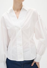 Blusa bianca con colletto e scollatura a V profonda, presenta maniche lunghe a sbuffo, chiusura anteriore con bottoni e una texture sottile. Adatta per varie occasioni.