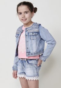 Denim jakna z dekorativnim šivanjem na ramenih, svetlo modre barve, v kombinaciji z roza topom z belim cvetličnim vzorcem in denimsko kratko hlačami z čipkastim robom.