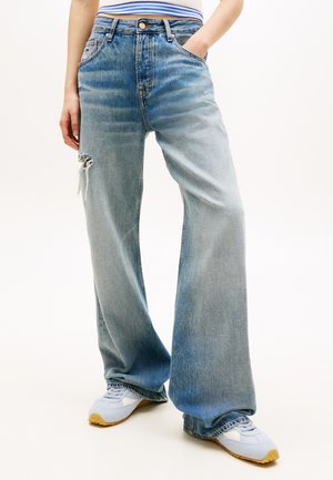 Persoon draagt loszittende, vervaagde blauwe spijkerbroek met een scheur aan de linker dij en witte sneakers met bruine zolen, staand met handen in de zakken.