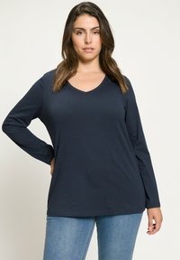 Námořnická modrá dlouhá rukávová halenka s výstřihem do V, vyrobena z měkké látky, uvolněného střihu, s rovým lemem a standardními manžetami, kombinována se světle modrými džíny.