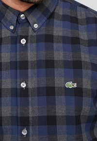 Hombre vistiendo una camisa de cuadros azul, gris y negro con el logotipo de cocodrilo verde bordado en el pecho.