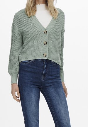 Mujer con cárdigan de punto verde claro con botones sobre camiseta blanca y vaqueros azul oscuro, de pie frente a fondo blanco.