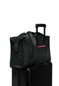 Sac de voyage noir en toile avec des doubles poignées, posé sur une valise rigide noire. Comprend une poche zippée rose et un détail de logo.