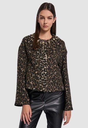 Donna che indossa una maglia a maniche lunghe con stampa leopardata e borchie metalliche vicino al collo e pantaloni in ecopelle neri, in piedi davanti a uno sfondo semplice.