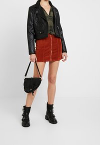 Schwarze Lederbikerjacke, olivgrünes gestreiftes Oberteil, rostfarbene Cordrock mit Frontreißverschluss, schwarze Knöchelstiefel und strukturierte Handtasche.