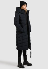 Geacă lungă neagră tip puffer cu guler înalt, cusături în formă de v și fermoar frontal, cu buzunare laterale. Asortată cu cizme negre până la gleznă și o căciulă tricotată.