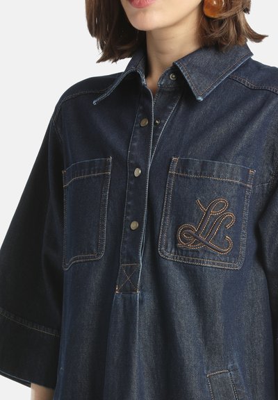 Denim-paita, jossa on kauluspää, kaksi rintataskua, napitus edessä, tummansininen väri ja kontrastivärinen ruskea brodeeraus yhdessä taskussa.