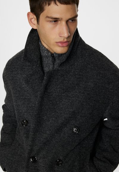 Cappotto grigio in lana doppiopetto con bottoni neri, colletto alto e strato interno grigio con zip. Texture liscia e design strutturato.