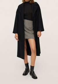 Lange zwarte jas met verlaagde schouders, een notch-kraag en een split. Gecombineerd met een aansluitend zwart topje en een rok met houndstooth-patroon. Zwarte laarzen.