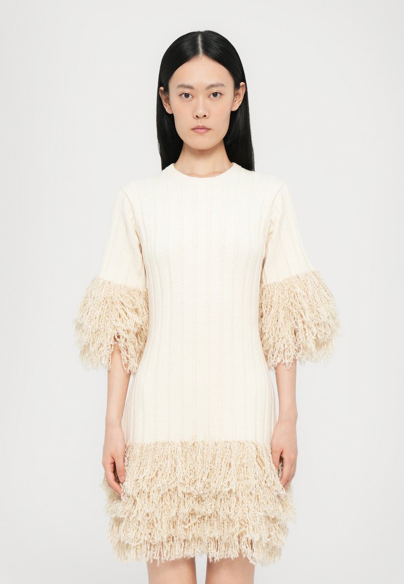 Robe en tricot écrue avec texture côtelée. Dotée de manches et d'ourlet frangés beiges et duveteux, créant un look superposé.