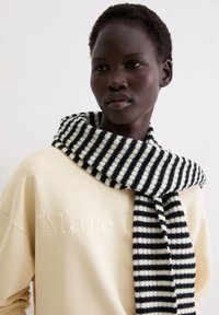 Czarnobiały prążkowany dziergany szalik zarzucony na szyj�ę, zestawiony z kremową bluzą z wypukłym logo.