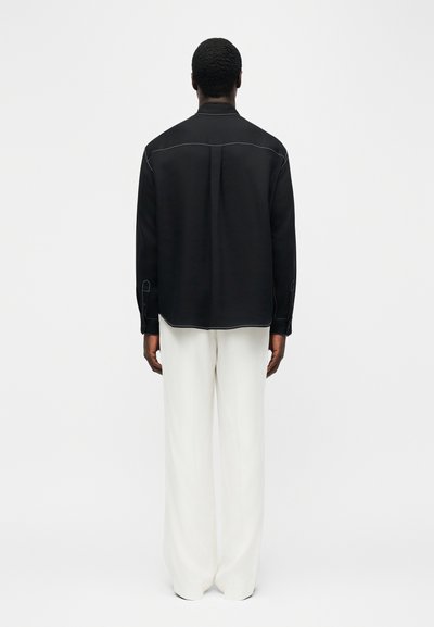 Άνδρας που φοράει μαύρο μακρυμάνικο πουκάμισο με λευκές ραφές και χαλαρό λευκό παντελόνι, στέκεται μπροστά σε απλό ανοιχτό γκρι φόντο.