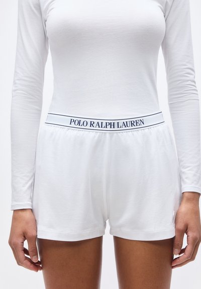 Λευκά σορτς με ελαστική μέση και λογότυπο "Polo Ralph Lauren", φορεμένα με μακρυμάνικη λευκή μπλούζα, που δείχνουν τον κορμό και τα πάνω πόδια.