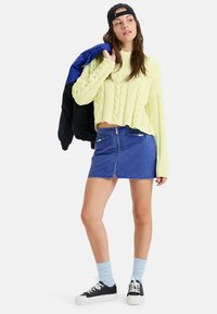 Junge Frau in einem gelben Zopfstrickpullover, blauer Jeans-Minikleid, schwarzer, rückwärts getragener Baseballmütze, die eine blau-schwarze Jacke über der Schulter trägt.