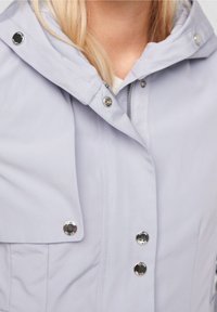 Veste gris clair avec une grande capuche, des boutons à l'avant et une fermeture éclair. Le matériau est lisse avec un léger éclat. Coutures détaillées visibles.