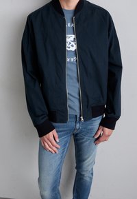 Chaqueta bomber azul marino con puños y cuello acanalados, cremallera frontal y tela ligera, usada sobre una camiseta gráfica gris y jeans azules.