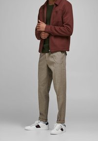 Bordowa kurtka na zamek, pod spodem zielony sweter z dzianiny, beżowe spodnie w kratę z łańcuszkowym detalem oraz białe sneakersy z kolorowymi paskami.