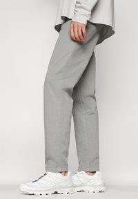 Personne portant un pantalon gris clair et des chaussures de sport blanches avec des logos colorés, debout devant un fond clair uni.