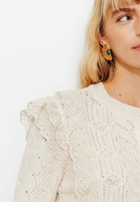 Pull en crème tricoté avec des motifs texturés, des détails en dentelle sur les épaules et un col côtelé. Le modèle porte de grandes boucles d'oreilles dorées ornées de pierres vertes.