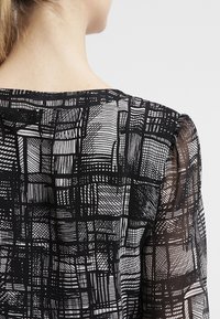 Blusa a pattern bianco e nero caratterizzata da un tessuto trasparente con design testurizzati a griglia e maniche a sbuffo.