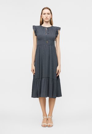 Kobieta stojąca w granatowej sukience midi w białe kropki, z rozkloszowanymi rękawami i białymi szpilkami na prostym tle.