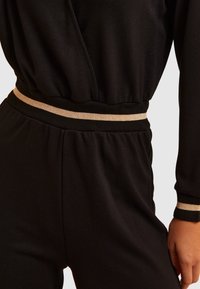 Nahaufnahme einer Person, die schwarze Hosen und ein Oberteil mit goldfarbenem und schwarzem elastischen Bund sowie Bündchen trägt, während die Hand an der Seite ruht.