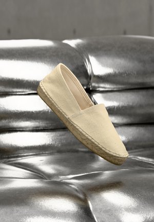 Beige lerret espadrillosko med flettet såle som svever over en blank, polstret overflate i sølvskinn.