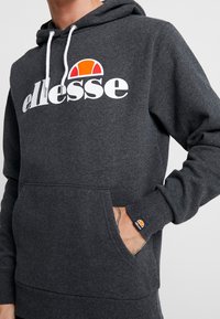 Felpa con cappuccio grigia caratterizzata da una tasca frontale, polsini a coste e un grande logo bianco "ellesse" con un accento arancione e giallo sopra di esso.