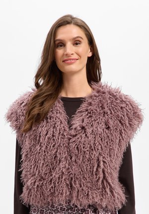 Jeune femme aux longs cheveux bruns porte un gilet mauve duveteux à texture bouclée sur un haut noir, souriant doucement sur un fond uni.