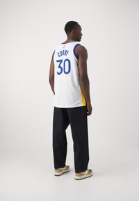 Basketbalový dres v bílé barvě s modrými a žlutými akcenty. Na zadní straně nese jméno "Curry" a číslo "30". Nosen s tmavými kalhotami a teniskami.