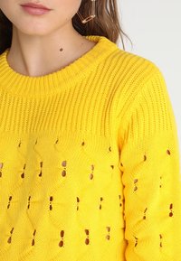Pull jaune en tricot avec un col rond côtelé et un motif ajouré, présentant des trous distincts et une surface texturée.