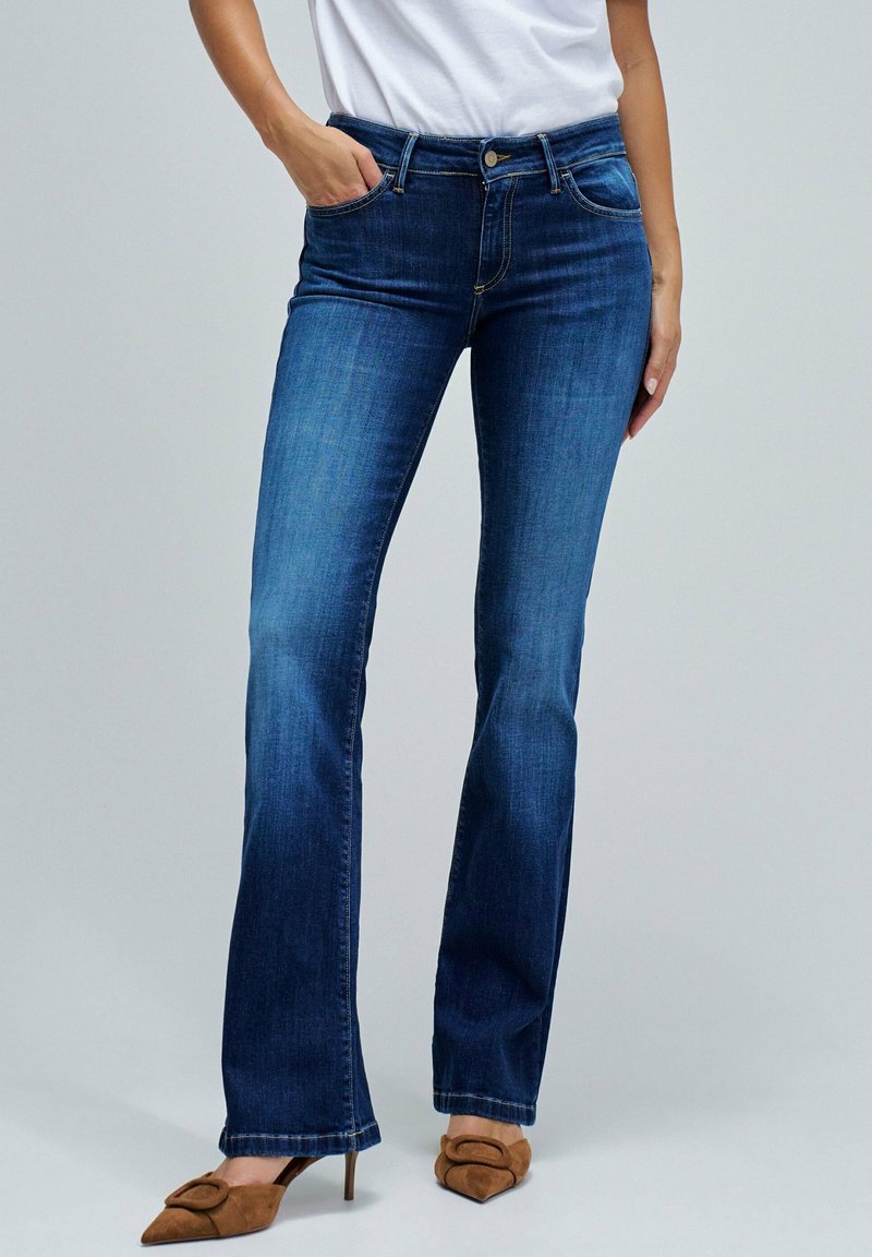 Mujer con jeans bootcut oscuros, camiseta blanca y zapatos de tacón altos de punta marrones, de pie con una mano en el bolsillo.